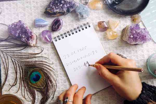 a lady using a pencil working out her numerology number. She has quartz crystals and a peacock feather surrounding the pad she is writing on